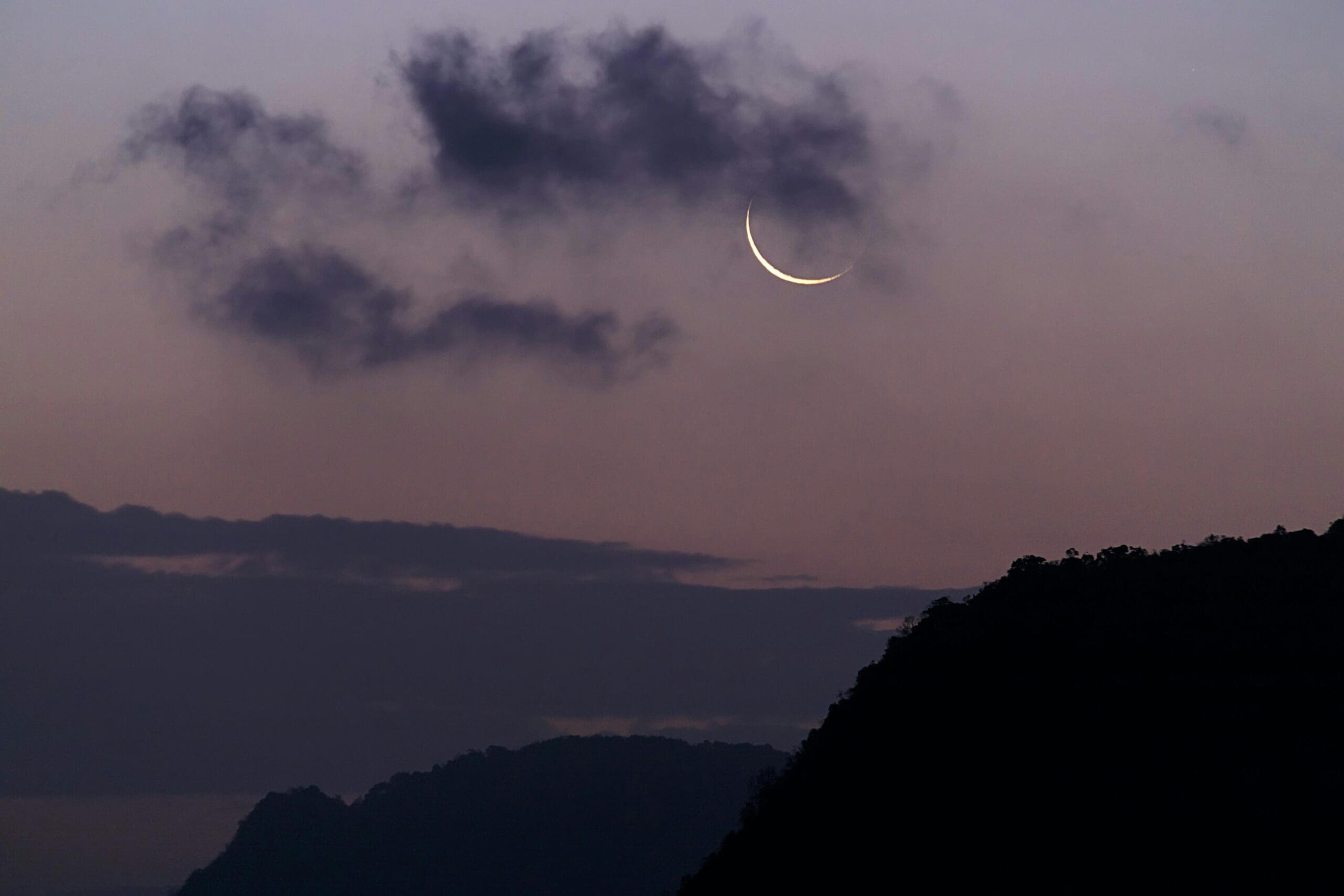 A purple sunset with the new crescent moon in the sky with clouds